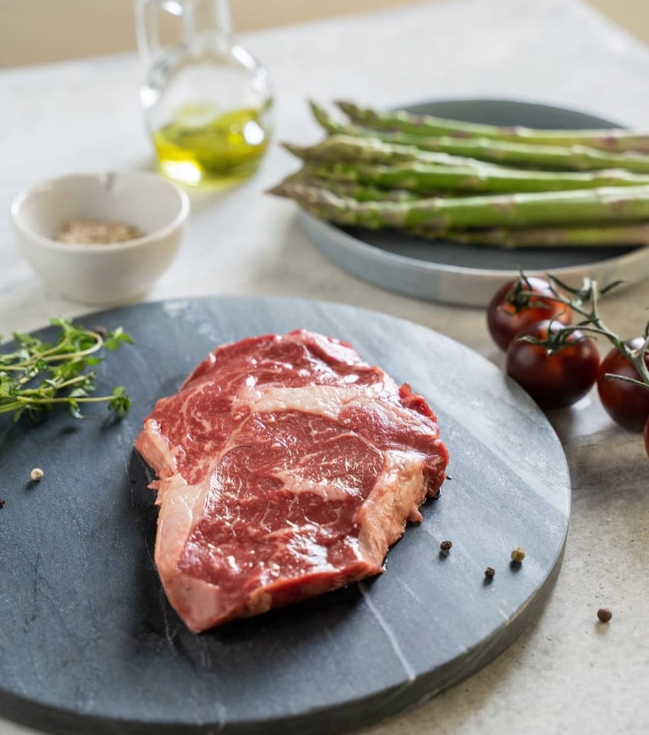 Raw ribeye steak on dark marble with asparagus, tomatoes and olive oil