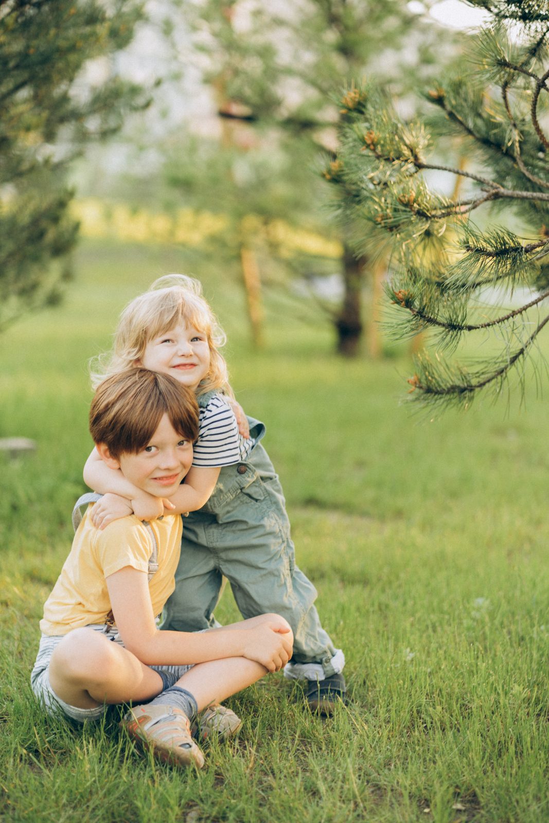 Little sister hugging her brother on the grass under pine trees