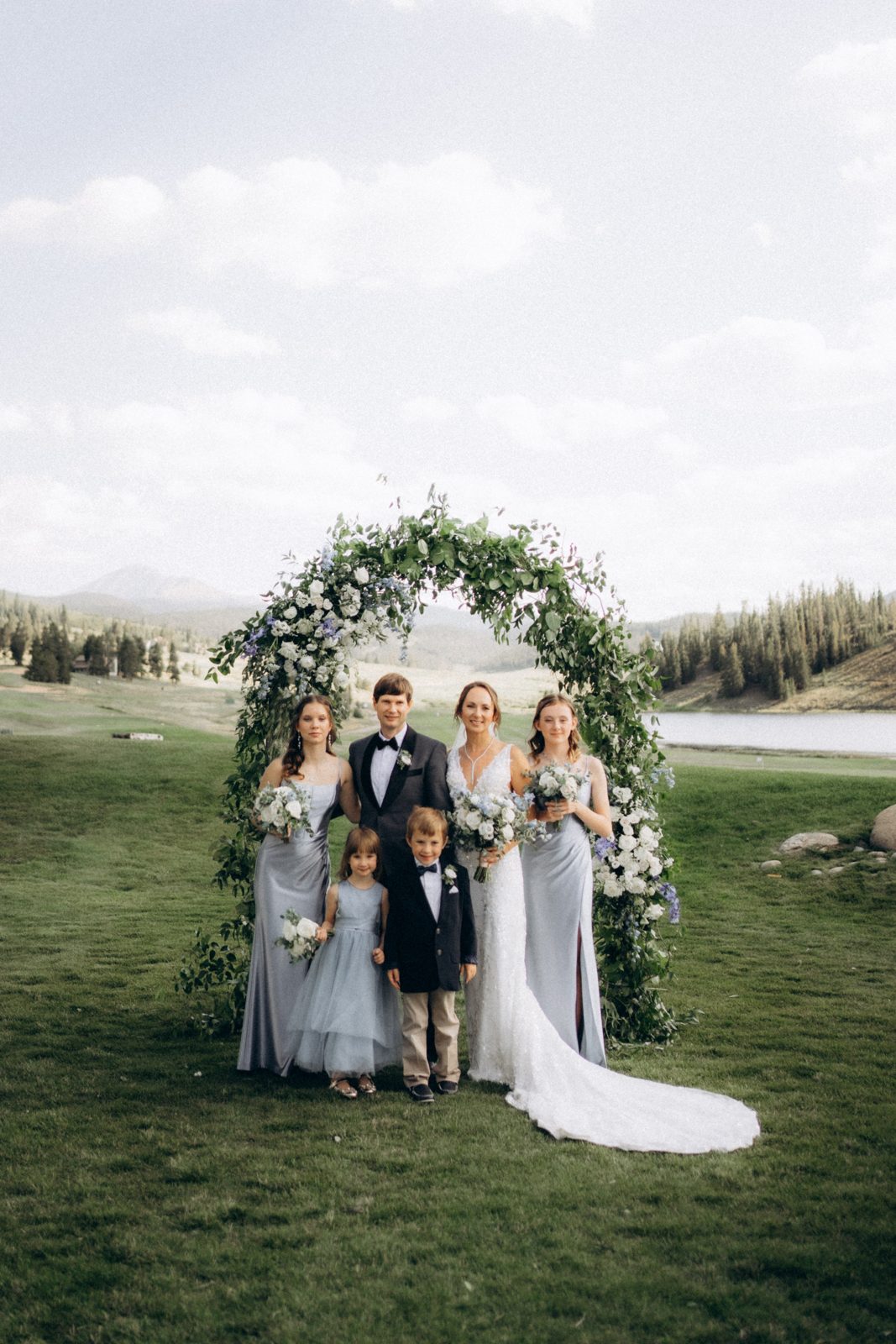 Bride, groom, bridesmaids and children under a floral arch by a mountain lake